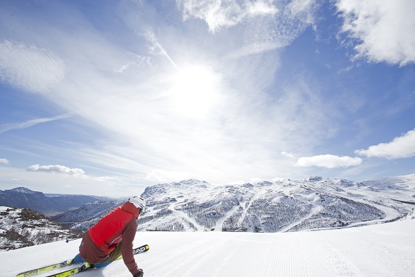 Person som åker skidor i en skidbacke. Snötäckta backar, blå himmel och sol.